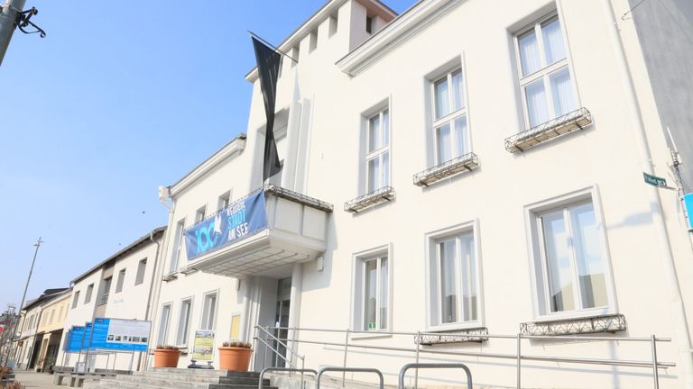 Ein weißes Gebäude mit mehreren Fenstern und einem blauen Banner, das '100 Jahre Stadt am See' steht. Eine schwarze Fahne hängt vom Balkon des Gebäudes.