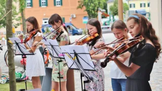 Fünf junge Mädchen spielen Geigen im Freien. Sie haben Geigenständer mit Notenblättern vor sich. Die Mädchen sind in Blumenkleidern gekleidet. Im Hintergrund befindet sich ein blauer Van und ein Gebäude.
