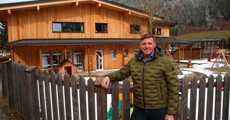A smiling man in a green jacket stands outside a wooden house with a fence in a snowy area.