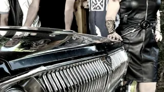 Vier Männer und eine Frau stehen hinter einem klassischen Auto. Die Frau trägt ein schwarzes Lederkleid. Sie alle schauen zur Kamera.