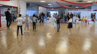 Eine Gruppe von Kindern führt einen Tanz in einem Saal mit Holzboden auf. Zuschauer sitzen im Hintergrund. Die Kinder tragen nummerierte Hemden.