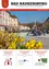 Das Bild zeigt einen Stadtplatz mit Menschen auf Bänken, einer Statue und Gebäuden mit Hinweisschildern für April 2026 und einer deutschen Webseite.