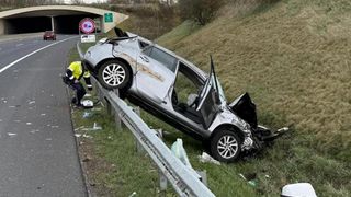 Ein Auto ist in eine Leitplanke geprallt und liegt nun auf dem Gras. Eine Person in einer gelben Weste untersucht die Unfallstelle.