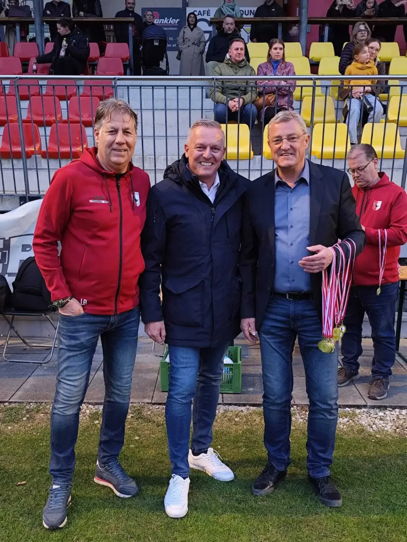 Drei Männer stehen auf einem Rasenfeld vor einem Stadion. Sie tragen Jacken und halten Medaillen. Dahinter sind Reihen leerer roter Sitze und ein Metallzaun.