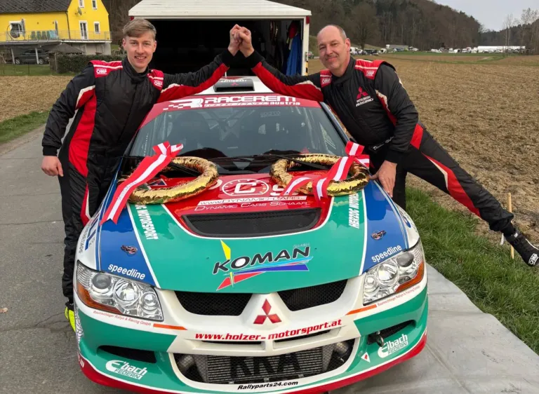 Zwei Männer in Rennanzügen stehen hinter einem grün-blauen Mitsubishi Lancer Evo, geschmückt mit einem Lorbeerkranz aus Goldblättern. Ein Mann hält einen goldenen Lorbeerkranz, während der andere seine Hand zum Siegspose erhoben hat.