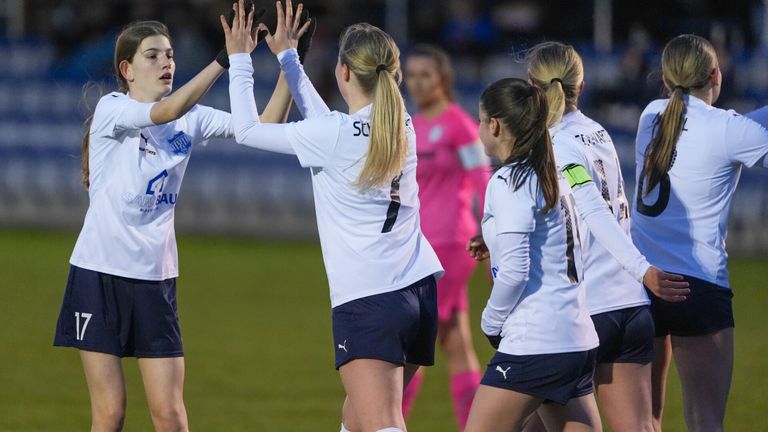 Vier Fußballerinnen in weißen Trikots feiern gemeinsam auf einem Feld. Nummer 50 ist in der Mitte. Im Hintergrund ist eine weitere Spielerin in einem rosa Shirt zu sehen.