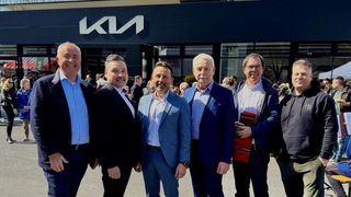 Fünf Männer in Anzügen stehen vor einem Gebäude mit einem großen weißen Buchstaben M. Sie lächeln und schauen zur Kamera. Einer hält ein rotes Buch.