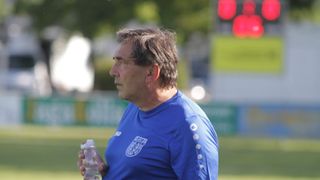 Ein älterer Mann in einem blauen Shirt mit einem Logo steht auf einem Feld und hält eine Flasche.