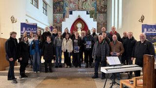 Ein Chor steht in einer Kirche mit Notenblättern, einige halten Bücher, einer spielt Klavier und andere posieren. Dahinter befinden sich religiöse Wandgemälde und ein Altar.