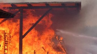 Ein Feuerwehrmann löscht mit einem Schlauch ein großes Feuer, das ein Gebäude eingenommen hat, wobei intensive Flammen sichtbar sind.