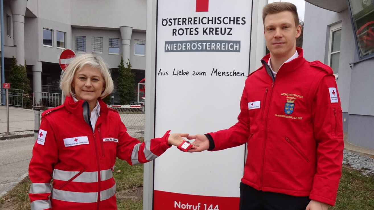 Ein Mann und eine Frau in roten Jacken stehen vor einem Schild des Österreichischen Roten Kreuzes und lächeln. Der Mann hält ein kleines Objekt in der Hand.