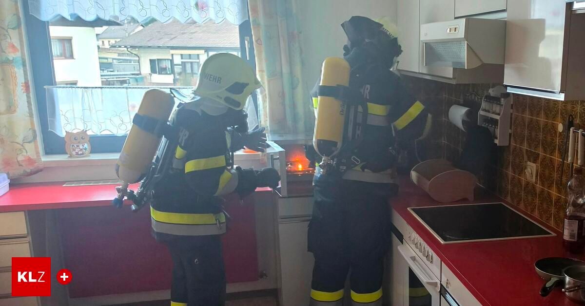 Zwei Feuerwehrleute in voller Schutzausrüstung stehen in einer Küche mit einer brennenden Kerze. Einer hält einen Feuerlöscher, der andere hat einen Sauerstofftank.