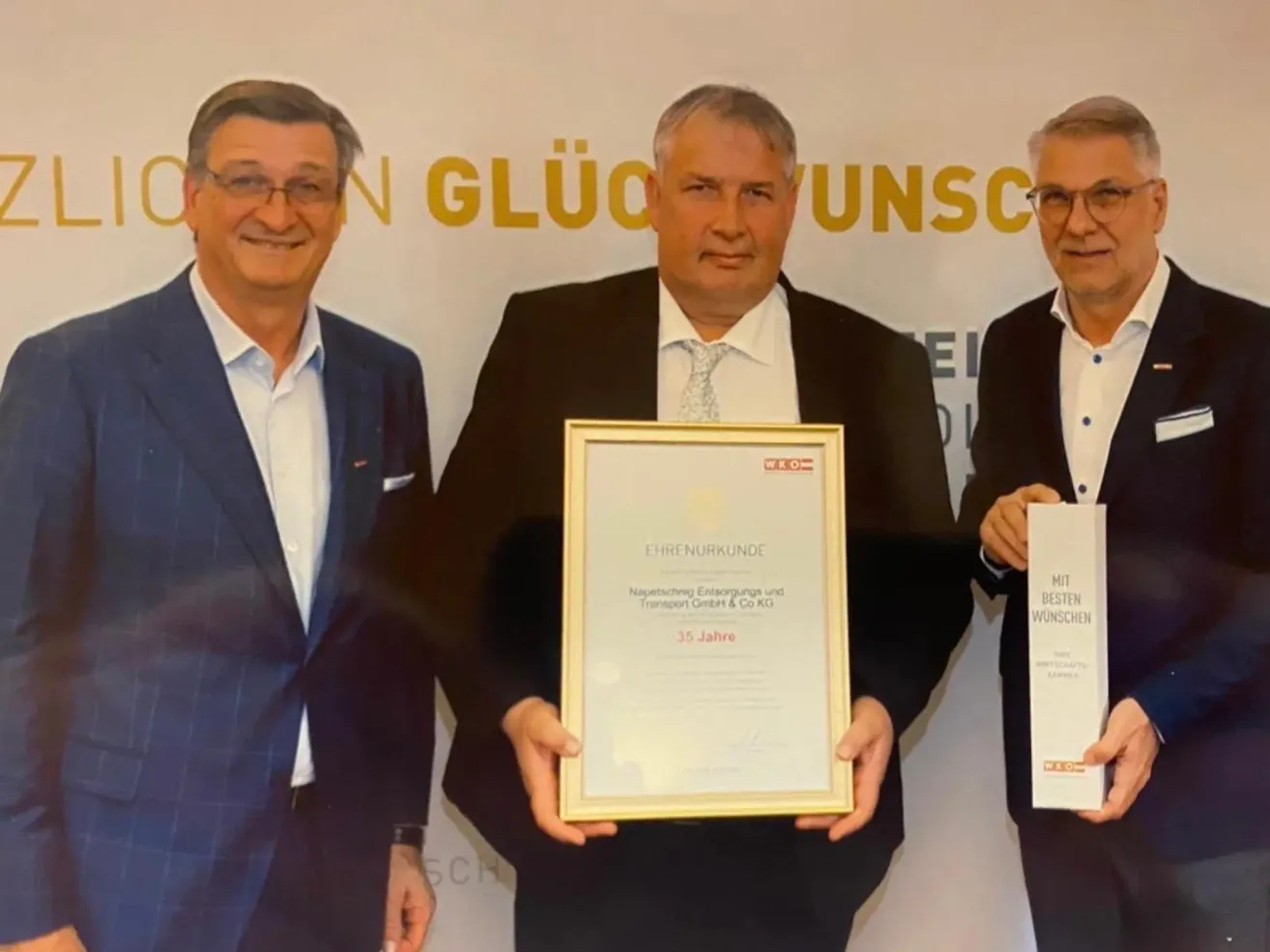 Drei Männer stehen vor einem Hintergrund mit goldenem Text. Der Mann in der Mitte hält ein gerahmtes Zertifikat. Die anderen halten Bücher. Sie tragen alle formelle Anzüge und Brillen.