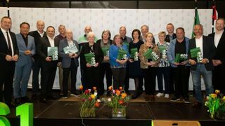 Eine Gruppe von Menschen mit grünen Büchern und Auszeichnungen steht in einer Reihe auf einer Bühne mit Blumen im Vordergrund.