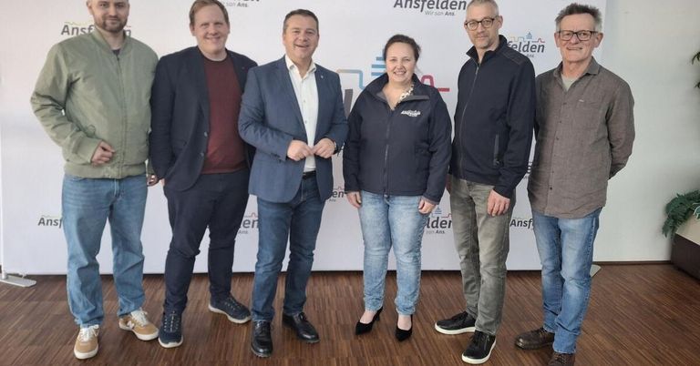 Eine Gruppe von Menschen in formeller und lässiger Kleidung steht vor einem weißen Hintergrund mit Logos und Text. Sie scheinen für ein Foto zu posieren.