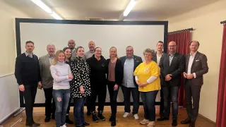 Eine Gruppe von Menschen steht vor einer Tafel in einem Raum, lächelt und posiert für ein Foto. Einige tragen Brillen und formelle Kleidung. Eine Frau in einem gelben T-Shirt sticht hervor. Der Raum hat Holzböden und rote Vorhänge.