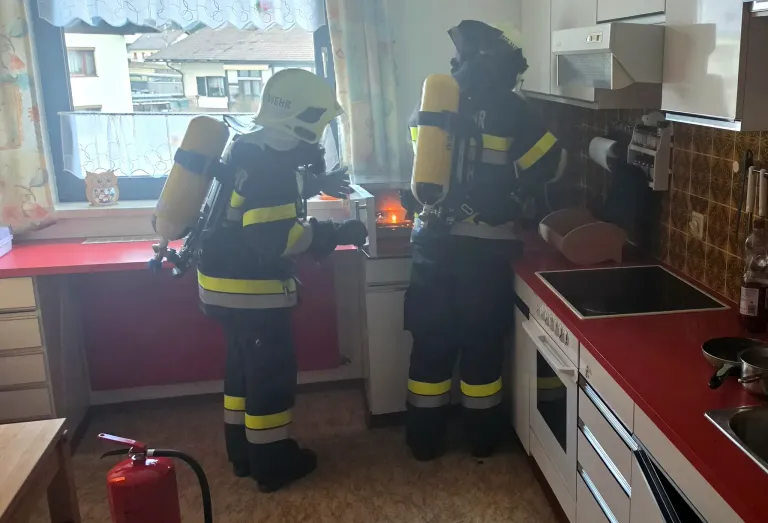 Zwei Feuerwehrleute mit Sauerstoffflaschen stehen in einer Küche mit einem Feuerlöscher und einem brennenden Feuer.