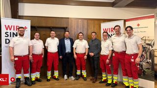 Eine Gruppe von Menschen in weißen und roten Uniformen, darunter zwei mit Brille, stehen vor einer Holzwand mit Bannern.