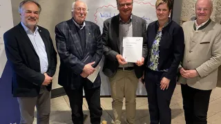 Fünf Menschen stehen in einem Raum. Ein Mann hält ein Zertifikat. Hinter ihnen sind Plakate und ein Whiteboard.