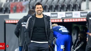 Ein Fußballtrainer mit Bart lächelt auf dem Feld, trägt eine schwarze Jacke. Hinter ihm sind zwei Personen anwesend, eine in einem blauen Trikot.