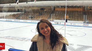 Eine Frau steht lächelnd vor einer Eisbahn. Dahinter ist die Bahn leer, einige Leute sitzen in den Tribünen. Werbung ist an der Barriere angebracht.