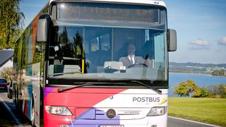 Ein Postbus ist in der Nähe eines Sees geparkt. Der Fahrer sitzt und trägt eine Krawatte. Der Bus ist lila, weiß und rot.