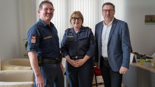 Drei Personen in Uniform stehen eng beieinander. Der Mann links trägt eine Brille und eine Dienstmarke. Die Frau in der Mitte lächelt. Der Mann rechts trägt einen Anzug.