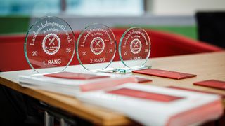 Drei Glaspreise auf einem Tisch, beschriftet mit 1., 2. und 3. Platz, mit dem Text 'Landeslehrlingswettbewerb' und '3. Lehrjahr'.