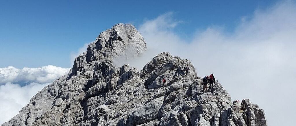 Bergsteiger erklimmen einen felsigen Berg unter einem klaren blauen Himmel. Nebel bedeckt teilweise den Gipfel des Berges.