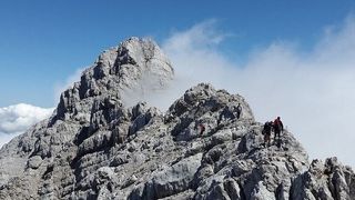 Bergsteiger erklimmen einen felsigen Berg unter einem klaren blauen Himmel. Nebel bedeckt teilweise den Gipfel des Berges.