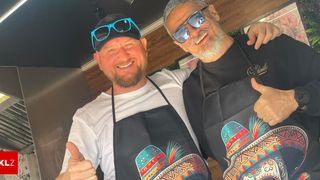 Zwei Männer in Schürzen lächeln und posieren für ein Foto. Der Mann auf der linken Seite trägt einen Hut mit Sonnenbrille.