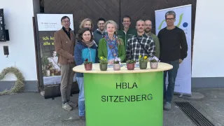 Eine Gruppe von Menschen steht hinter einem grünen Tresen mit der Aufschrift HBLa Sitzenberg, mit kleinen Topfpflanzen darauf. Sie sind lässig gekleidet und lächeln. Dahinter befinden sich zwei Banner mit Text und Bildern.