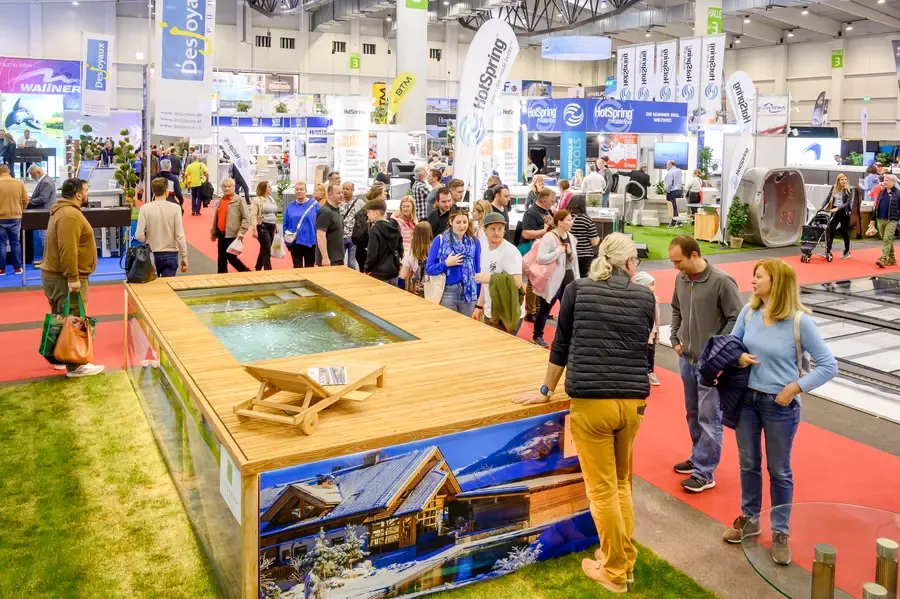 Besucher einer Messe mit einem Ausstellungsstück, das einen hölzernen Pool mit Wasser und einem Chalet-Design zeigt.