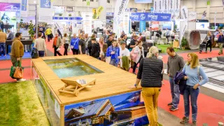Besucher einer Messe mit einem Ausstellungsstück, das einen hölzernen Pool mit Wasser und einem Chalet-Design zeigt.