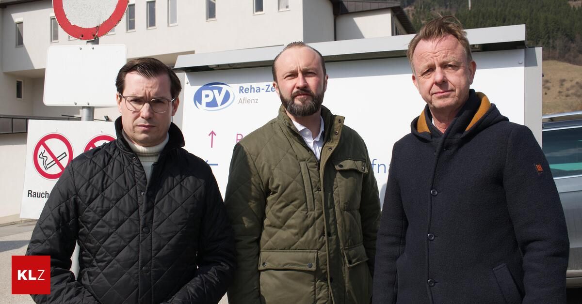 Drei Männer stehen vor einem Gebäude mit dem Schild 'Reha-Zentrum Aflenz'. Einer trägt eine Brille, der andere eine grüne Jacke und der dritte einen schwarzen Mantel. Sie sehen ernst aus.