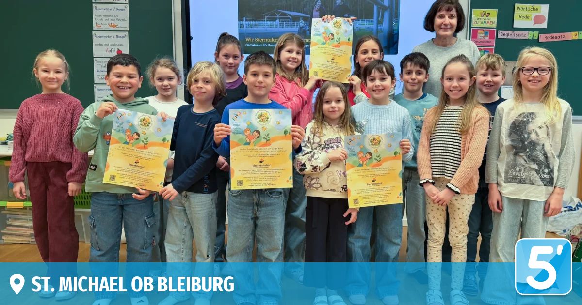 Eine Gruppe von Kindern im Klassenzimmer hält Urkunden mit strahlenden Gesichtern. Eine erwachsene Frau steht hinter ihnen, und eine Tafel befindet sich hinter den Kindern.