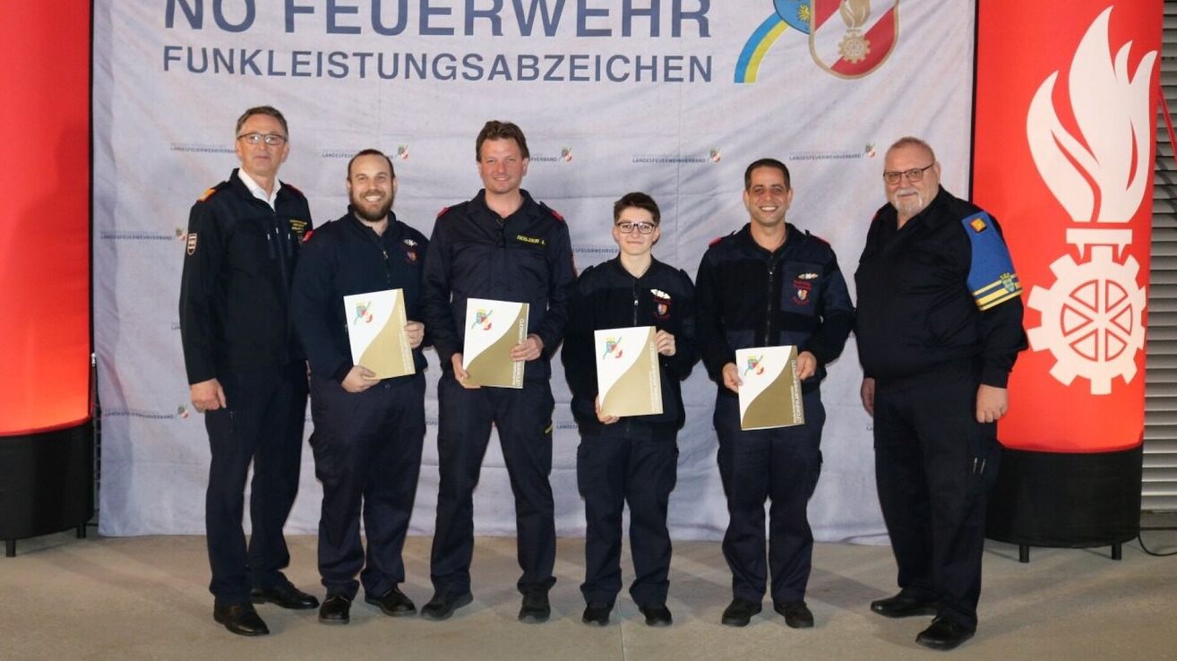 Fünf Feuerwehrleute in Uniform stehen vor einem Banner. Sie halten Zertifikate und posieren für ein Foto. Hinter ihnen ist ein weißes Banner mit einem Emblem.