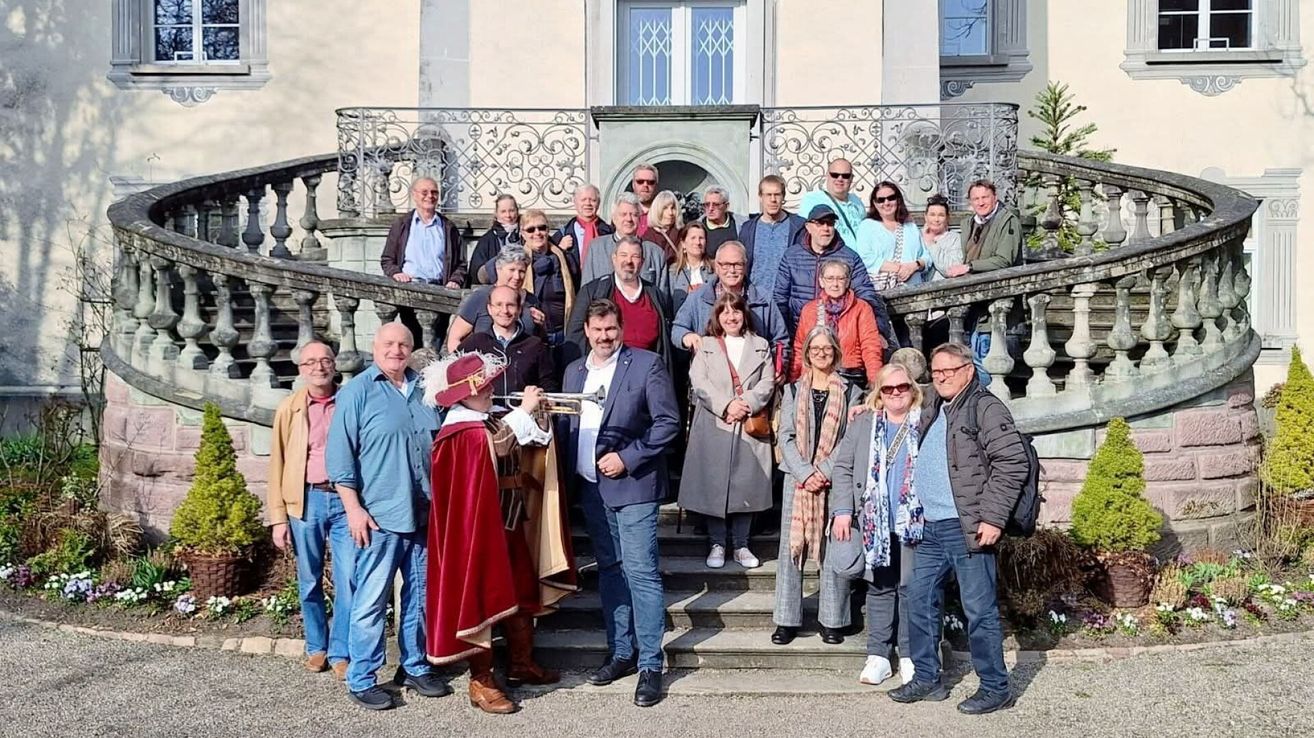 Eine Gruppe von Menschen posiert für ein Foto auf den Stufen eines historischen Gebäudes. Eine Person in einem Kostüm spielt Trompete.