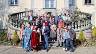 Eine Gruppe von Menschen posiert für ein Foto auf den Stufen eines historischen Gebäudes. Eine Person in einem Kostüm spielt Trompete.