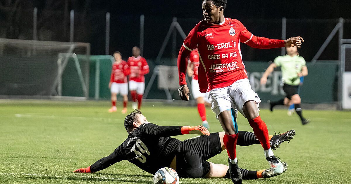Ein Fußballspieler in Rot taucht ab, um einen Schuss zu blockieren, während ein anderer Spieler in Schwarz fällt. Sie spielen bei Nacht mit verschwommenen Hintergrundspielern.