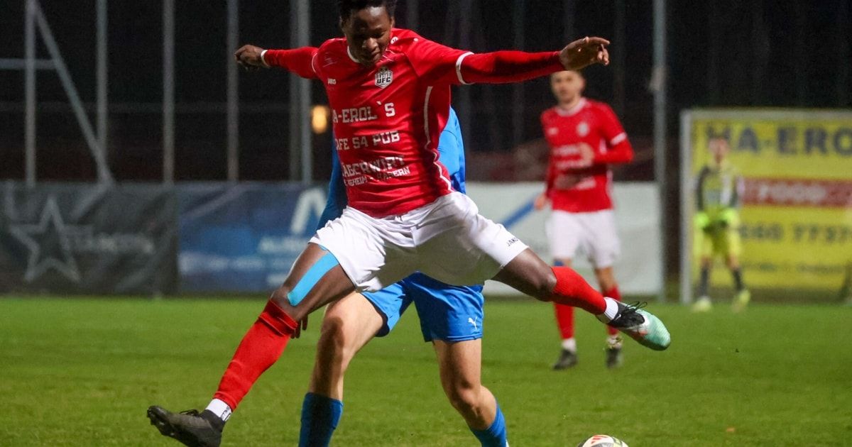 Fußballspieler in rotem Trikot tritt mit dem rechten Fuß an den Ball, während der Gegner versucht, den Schuss zu blockieren. Im Hintergrund läuft ein weiterer Spieler und zeigt das Feld.