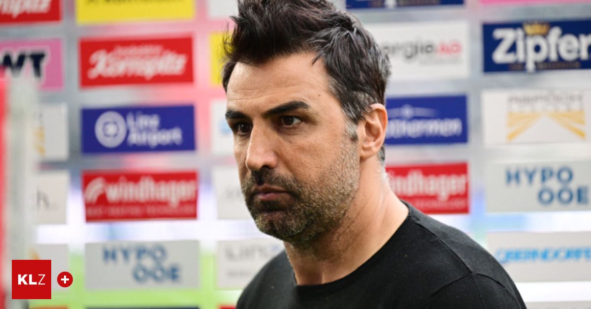 Ein Mann mit Bart und Oberlippenbart in einem schwarzen T-Shirt steht vor einer Wand mit Werbung. Er schaut zur Seite.