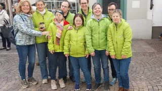 Eine Gruppe von Menschen in grünen Jacken posiert für ein Foto vor einem Gebäude. Eine Person hält eine Dose.