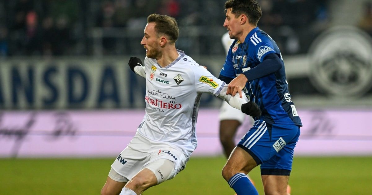 Zwei Fußballspieler sind auf dem Spielfeld in Aktion. Einer ist in Weiß, der andere in Blau, beide kämpfen erbittert.
