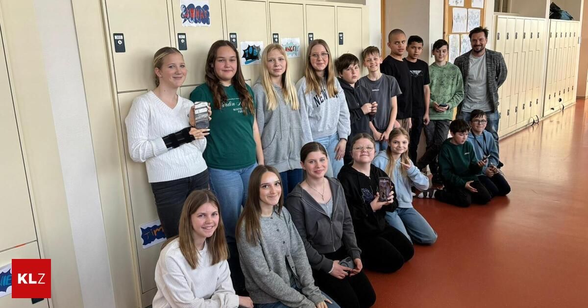 Eine Gruppe von Schülern, Jungen und Mädchen, steht und sitzt zusammen in einem Schulflur. Sie alle haben Lächeln im Gesicht und halten Auszeichnungen in den Händen. Schließfächer säumen die Wände hinter ihnen.