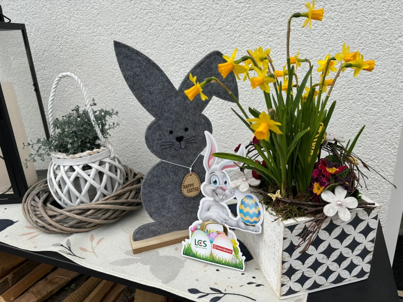 Ein dekorativer Osterhase hält ein Osterei in der Hand, neben einer Pflanze mit gelben Blüten in einem Korb. Ein Korb mit einer Pflanze steht hinter dem Hasen.