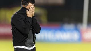 Ein Mann in einem schwarzen Jackett steht auf einem Feld und bedeckt sein Gesicht mit den Händen.