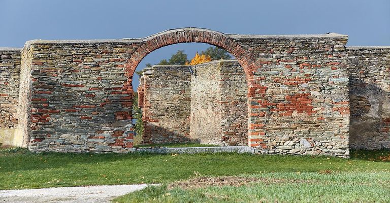 Ein alter Ziegelbogen steht in der Mitte einer Steinmauer, umgeben von Gras und Bäumen.