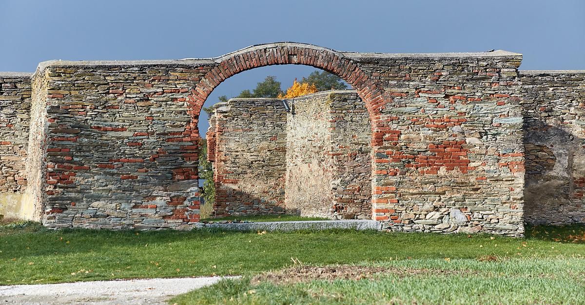 Ein alter Ziegelbogen steht in der Mitte einer Steinmauer, umgeben von Gras und Bäumen.