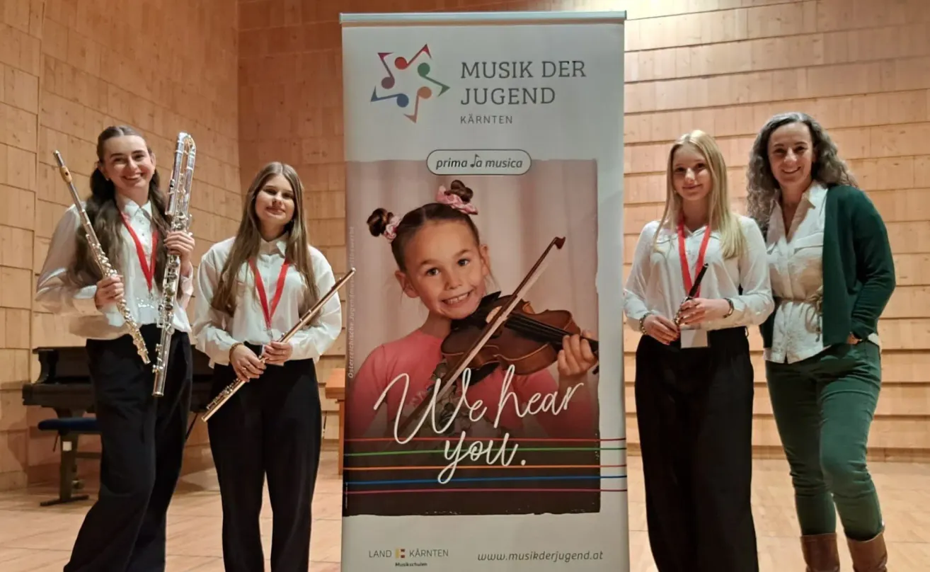 Drei Frauen stehen hinter einem Banner mit einem lächelnden Mädchen mit einer Geige, das ein Musikprogramm für Jugendliche bewirbt. Der Text lautet 'Wir hören Sie'.